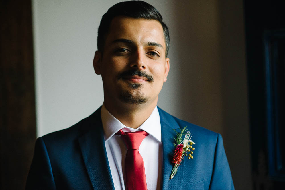Image showing an example of an indoor photography tip without flash of a groom in a blue suit with a red tie and a flower lapel before his wedding ceremony shot with window light