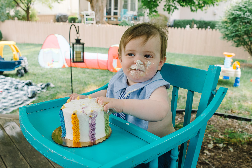 A young boy in a blue high chair with icing on his face and his hands in a small white birthday cake.
