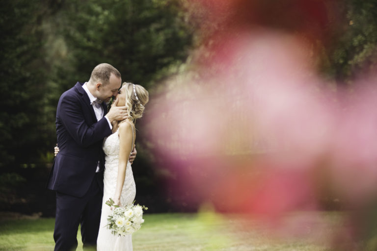 A bride and groom on their wedding day kissing with a red flower in the foreground.