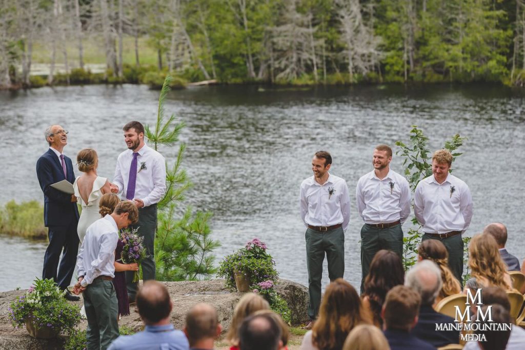 Image of a wedding on a lake of groomsmen look on as the bride and groom of the wedding exchange vows and share a funny moment