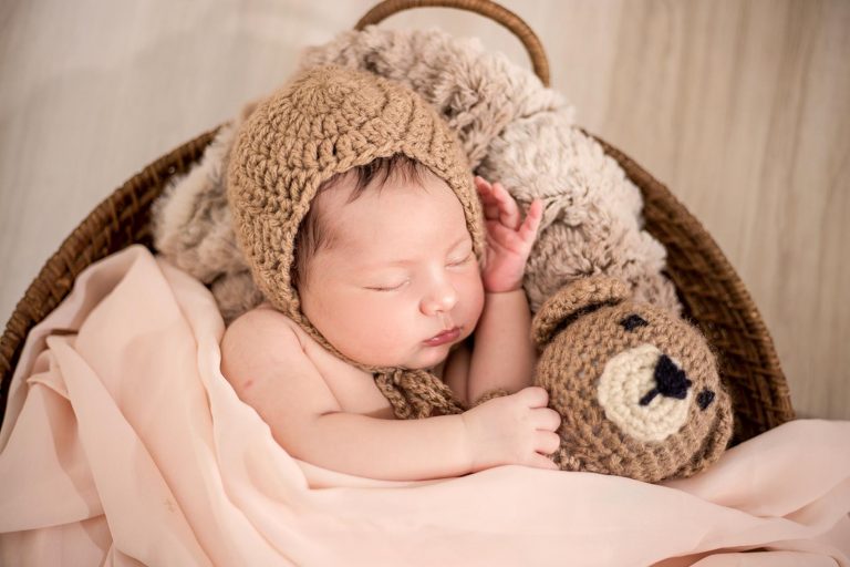 A newborn baby with a hat on sleeping in a basket with a stuffed animal bear.