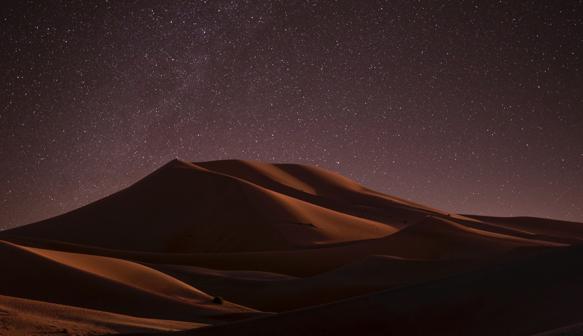 A sand dune in a desert with a night sky and stars in the background.