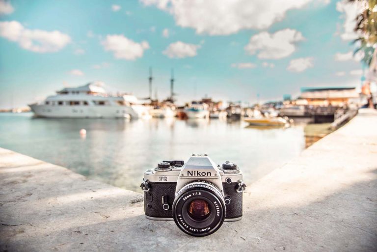 A silver Nikon film camera on a concrete ledge in front of a bay with boats and a blue sky with some white clouds.