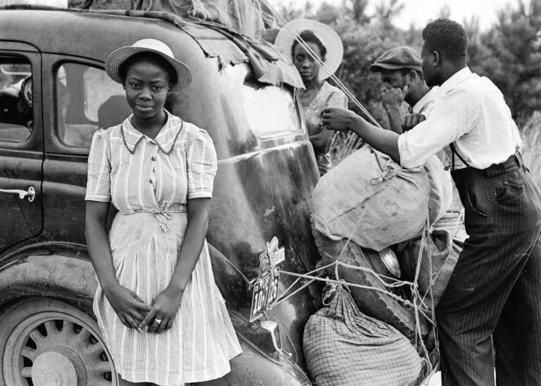 People loading stuff into a car with a woman looking into the camera in black and white.