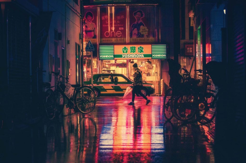 Urban photo of an alley with a woman with an umbrella walking in front of a store at night with a wet, reflective ground