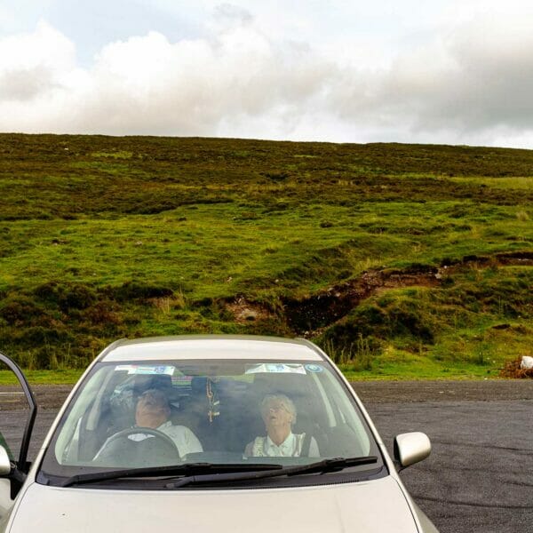 Old couple parked and asleep in a silver car in front of a lush green hill on a cloudy day.