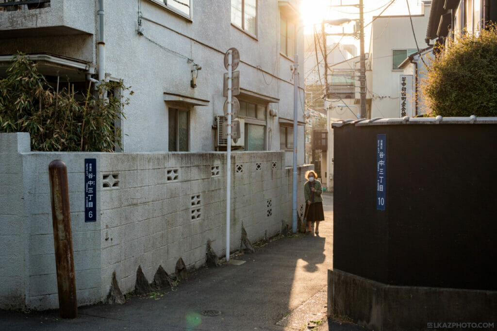 An old woman sweeping in an alley with the sun behind her.