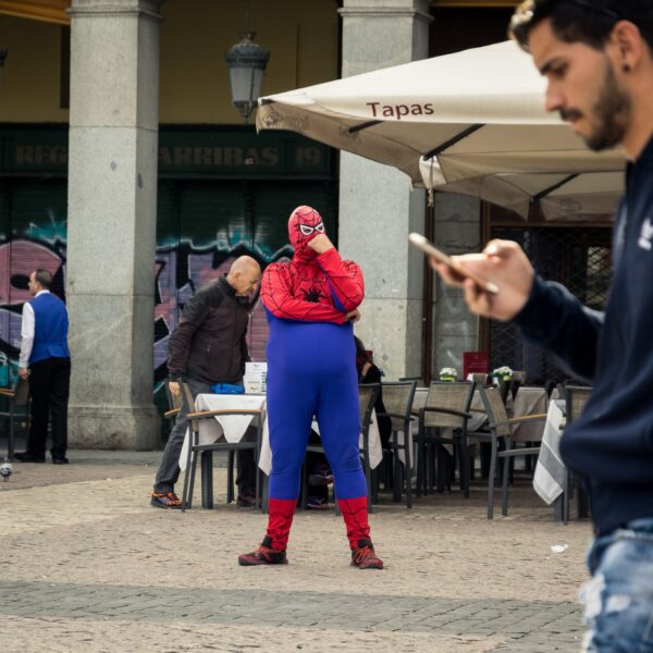 A person dressed as spider man in a public place with a man holding his phone walking in front.