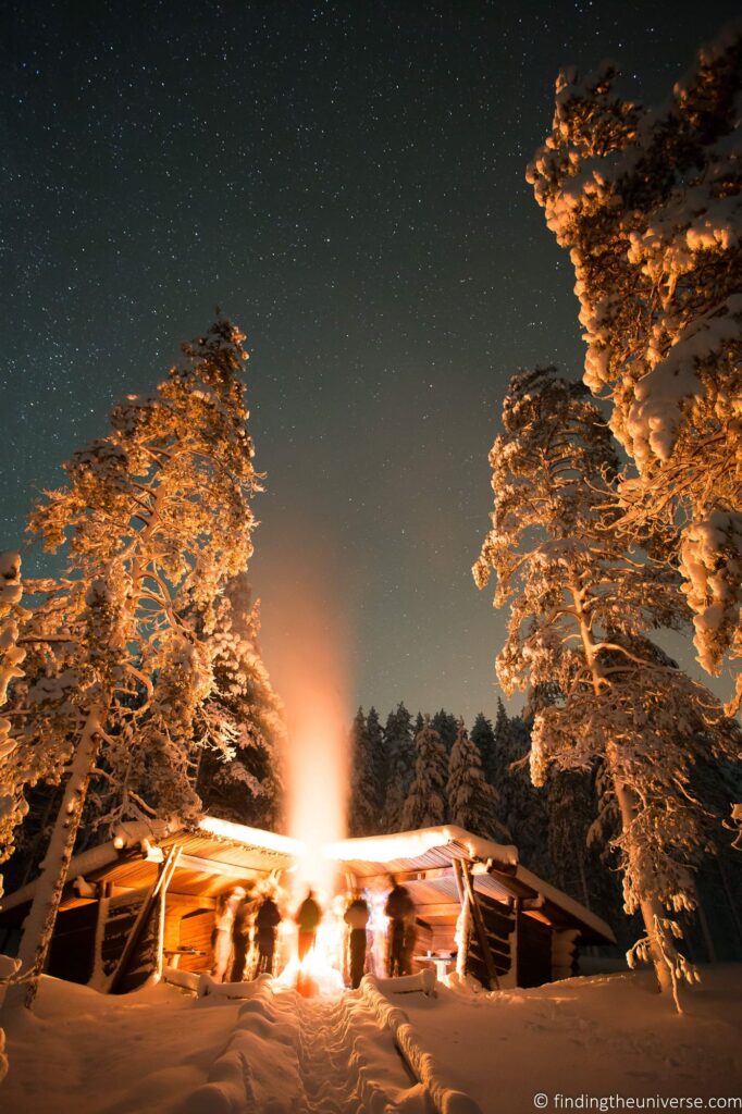 A group of people around a fire in a snowy place with tall fir trees.