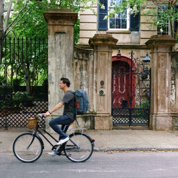A man wearing a backpack riding on a bike in front of an old building with a gate.