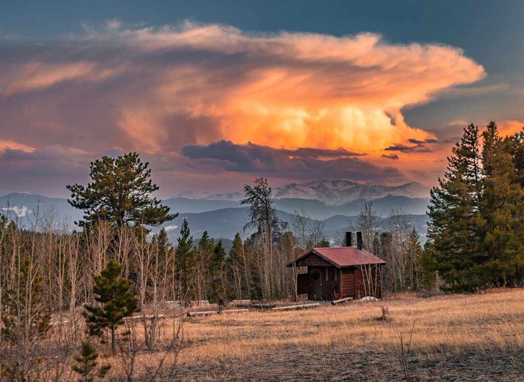 A landscape with a dramatic sky at golden hour with a red barn and mountains in the background.